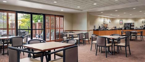 A hotel dining area with tables and chairs, a counter with a coffee machine, and large windows with a view of greenery outside.