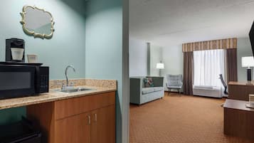 A hotel room with a kitchenette, a sofa, a chair, a desk, and a large window with curtains.