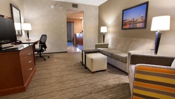 Quarto de hotel com escrivaninha, cadeira de escritório, sofá, banquinho e televisão.
