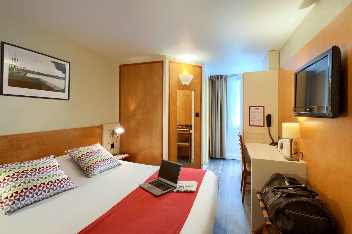Una habitación de hotel con una cama, una computadora portátil, un escritorio con lámpara, un televisor y una maleta.
