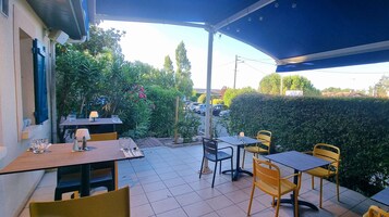 Un patio al aire libre con mesas y sillas, un área techada y vista a coches estacionados y follaje.