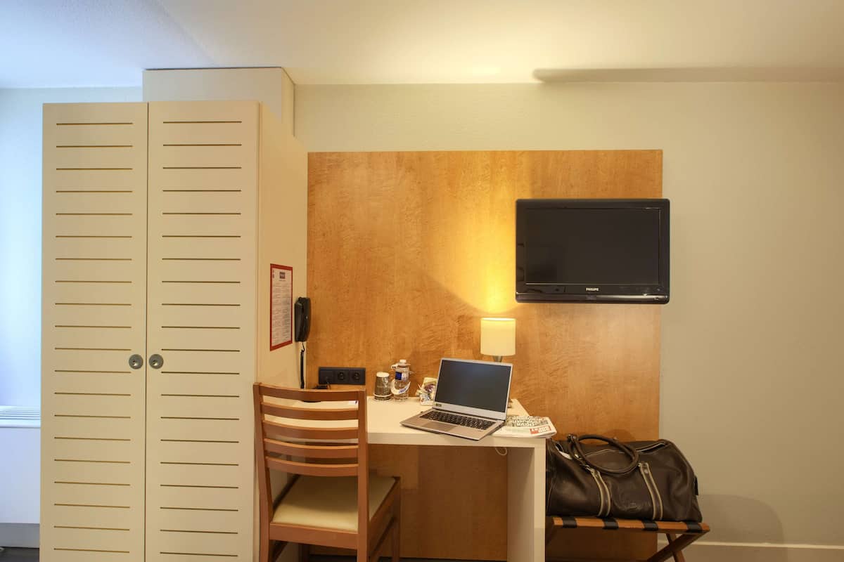A compact hotel room with a desk, a laptop, a television mounted on the wall, a chair, and a wardrobe.