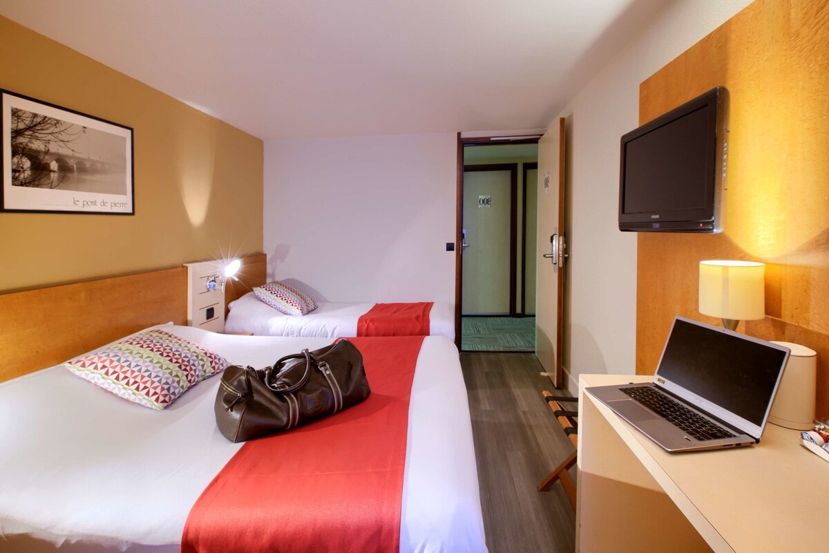 Una habitación de hotel con dos camas, un escritorio con una computadora portátil, un televisor y una puerta que da a otra habitación.