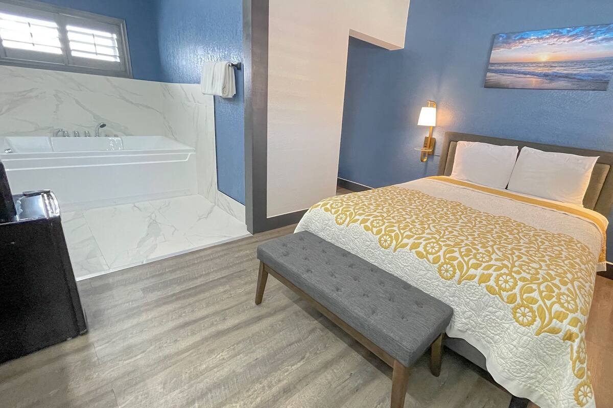 A bedroom with a bed, bedside lamps, a bench, a painting, and a bathroom with a bathtub.