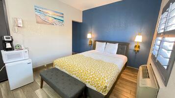 A hotel room with a bed, bedside lamps, a painting of a beach, a microwave, and a bench.