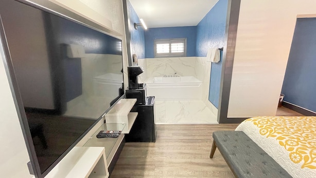 A modern hotel room with a large flat-screen TV, a small desk with a coffee maker, a bathtub, and a bed with a patterned bedspread.