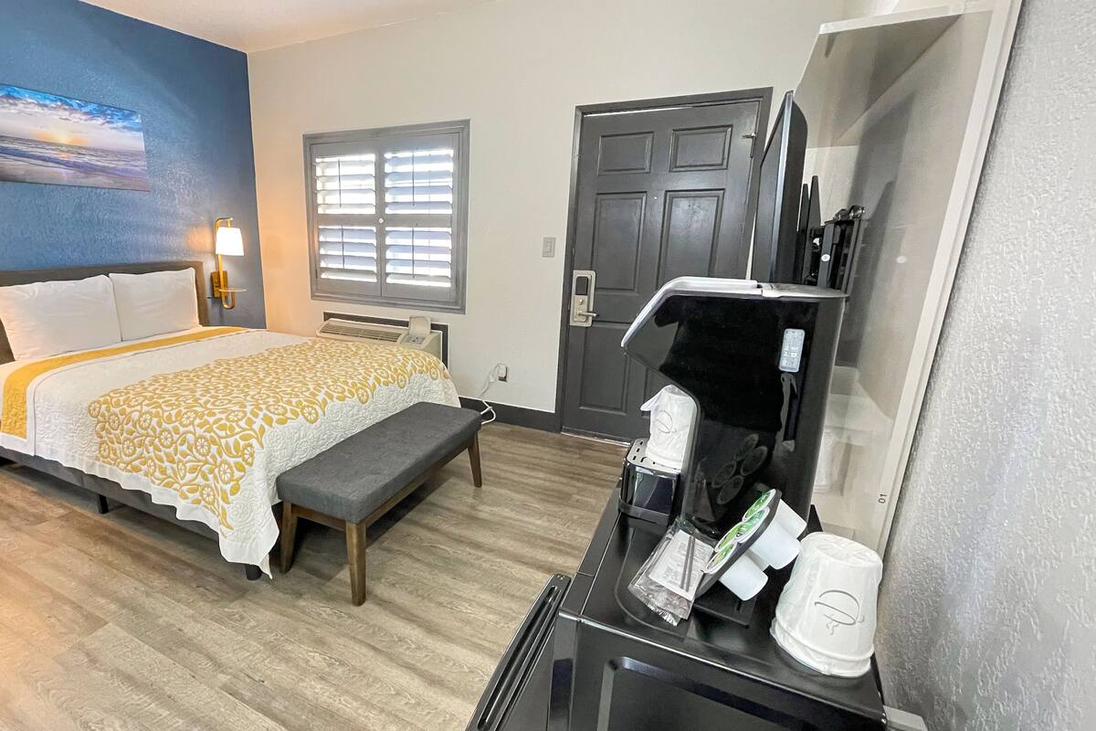 A hotel room with a bed, bench, bedside lamps, a window with blinds, a door, and a coffee machine.