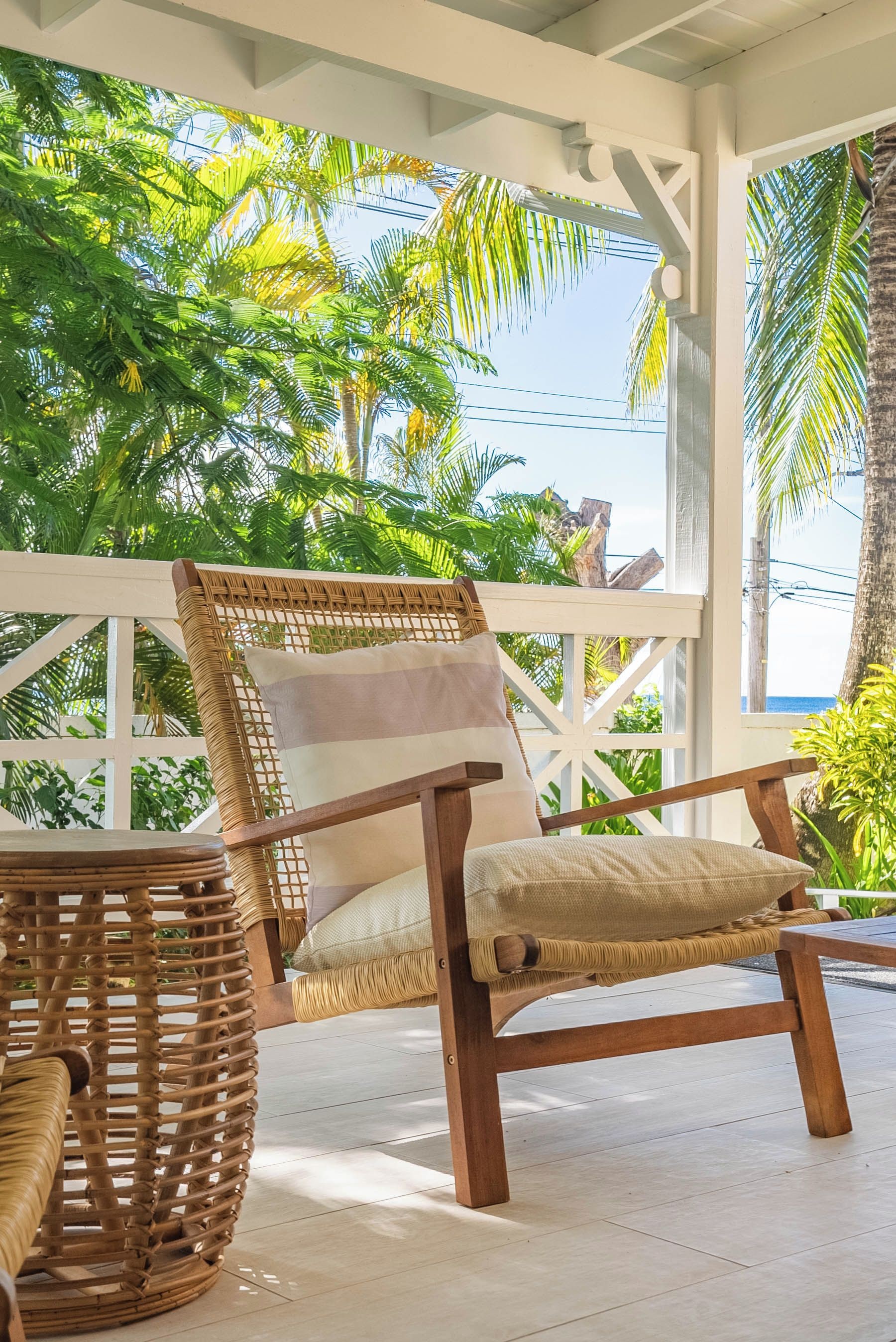 A wicker chair with a cushion and a woven basket, set on a tiled floor under a white ceiling structure with a view of palm trees and the sea.