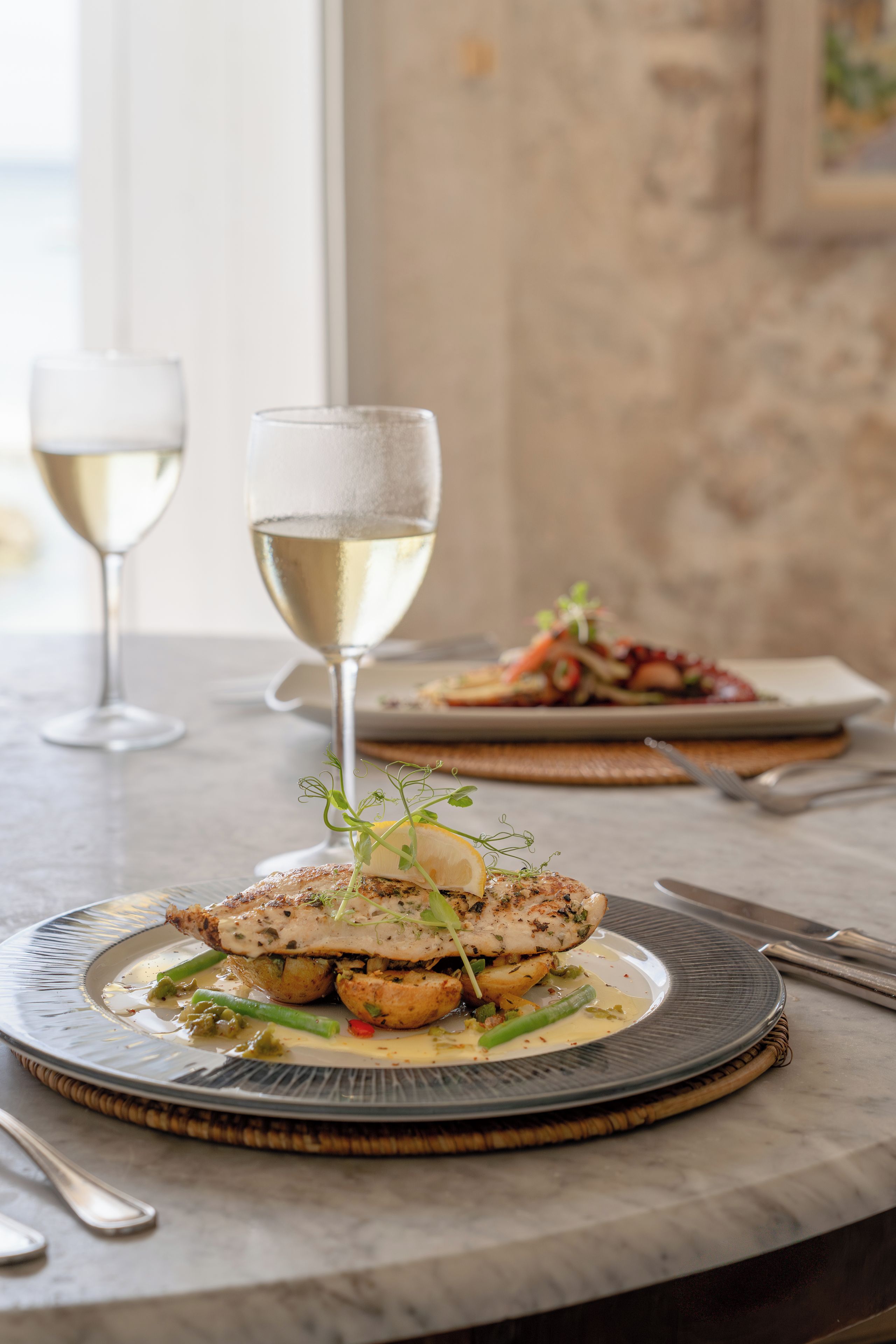 Una mesa de comedor con platos de comida y dos copas de vino blanco.