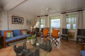 A living room with a blue sofa, orange cushions, a glass coffee table, and a view of the sea. - Little Good Harbour (Speightstown)