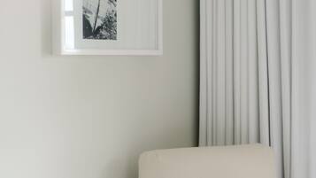 A white armchair with a patterned pillow, a framed black and white photograph on the wall, and a light-colored curtain.