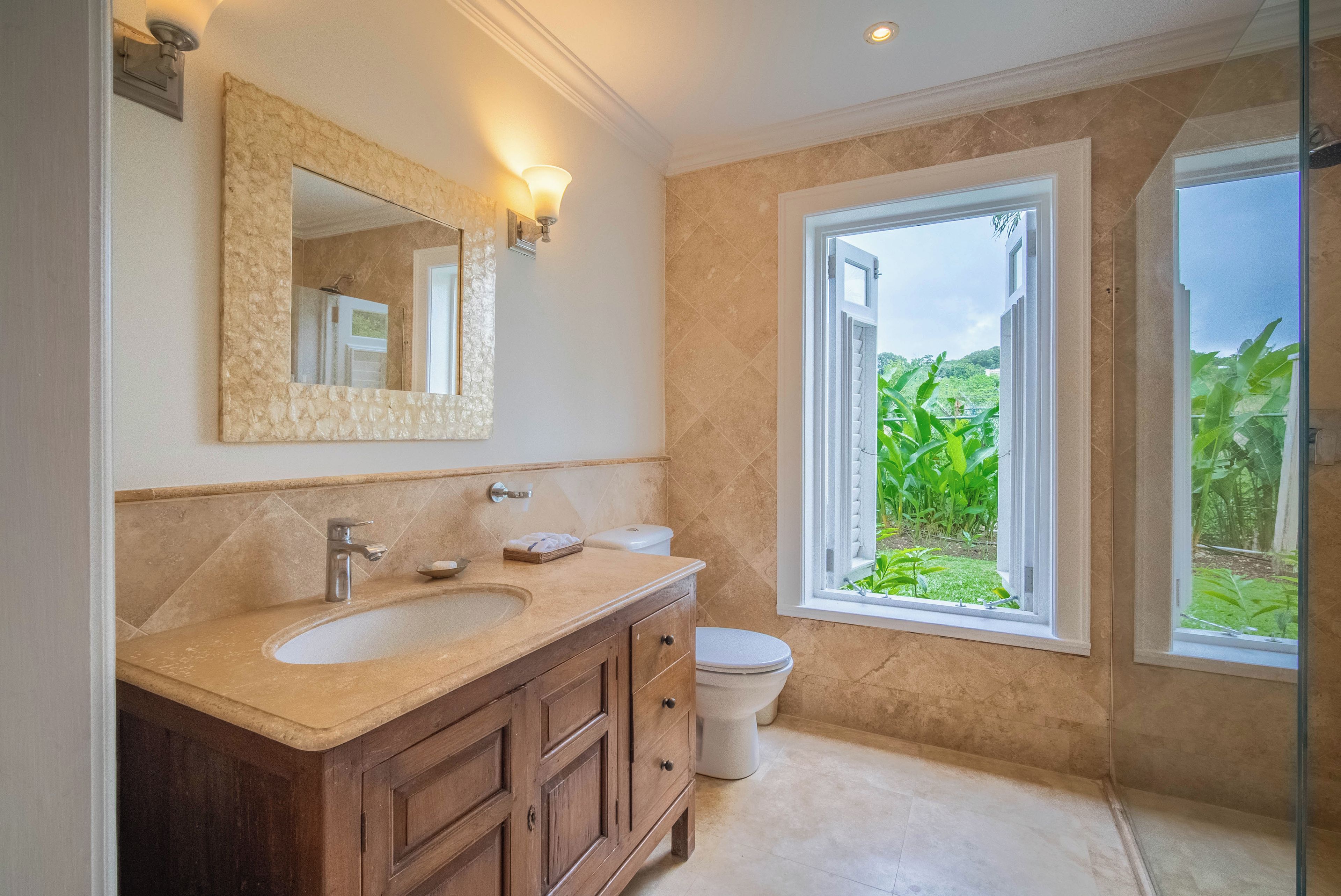 A bathroom with a double sink, a toilet, a large mirror, and a window with a view of greenery.