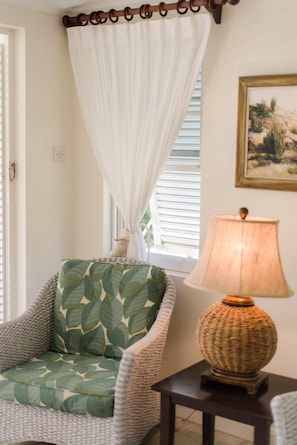 A wicker chair with a leaf-patterned cushion, a table with a woven lamp, and a window with white curtains. - Little Good Harbour (Speightstown)