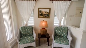 A room with two wicker chairs with leaf-patterned cushions, a small table with a lamp, and a painting on the wall. - Little Good Harbour (Speightstown)