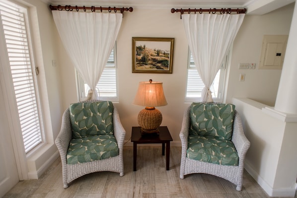 A room with two wicker chairs with leaf-patterned cushions, a small table with a lamp, and a painting on the wall. - Little Good Harbour (Speightstown)