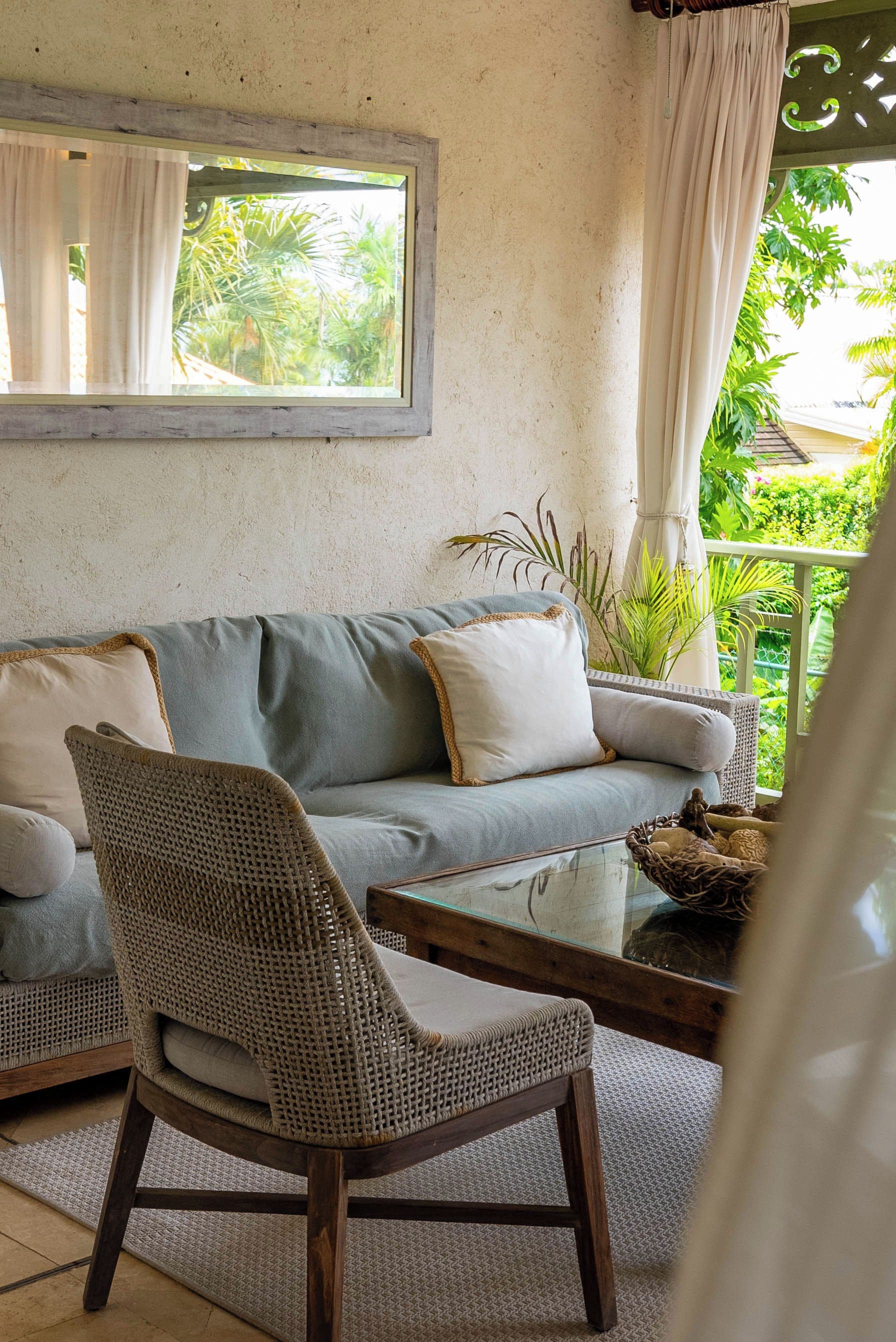 A cozy living area with a sofa, a wicker chair, a glass coffee table, and a view of greenery outside.