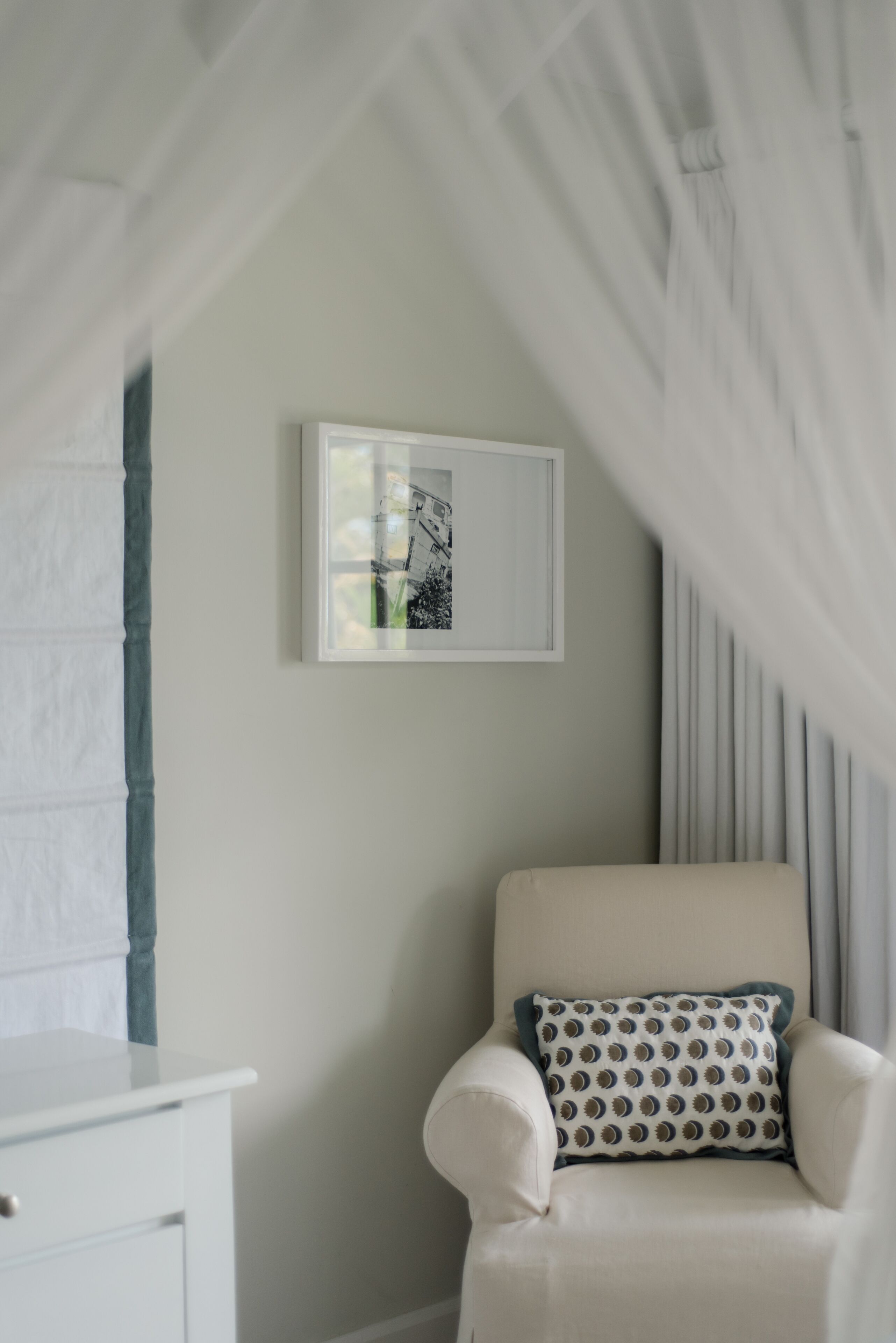 A room with a white armchair, a framed picture on the wall, and a sheer curtain.