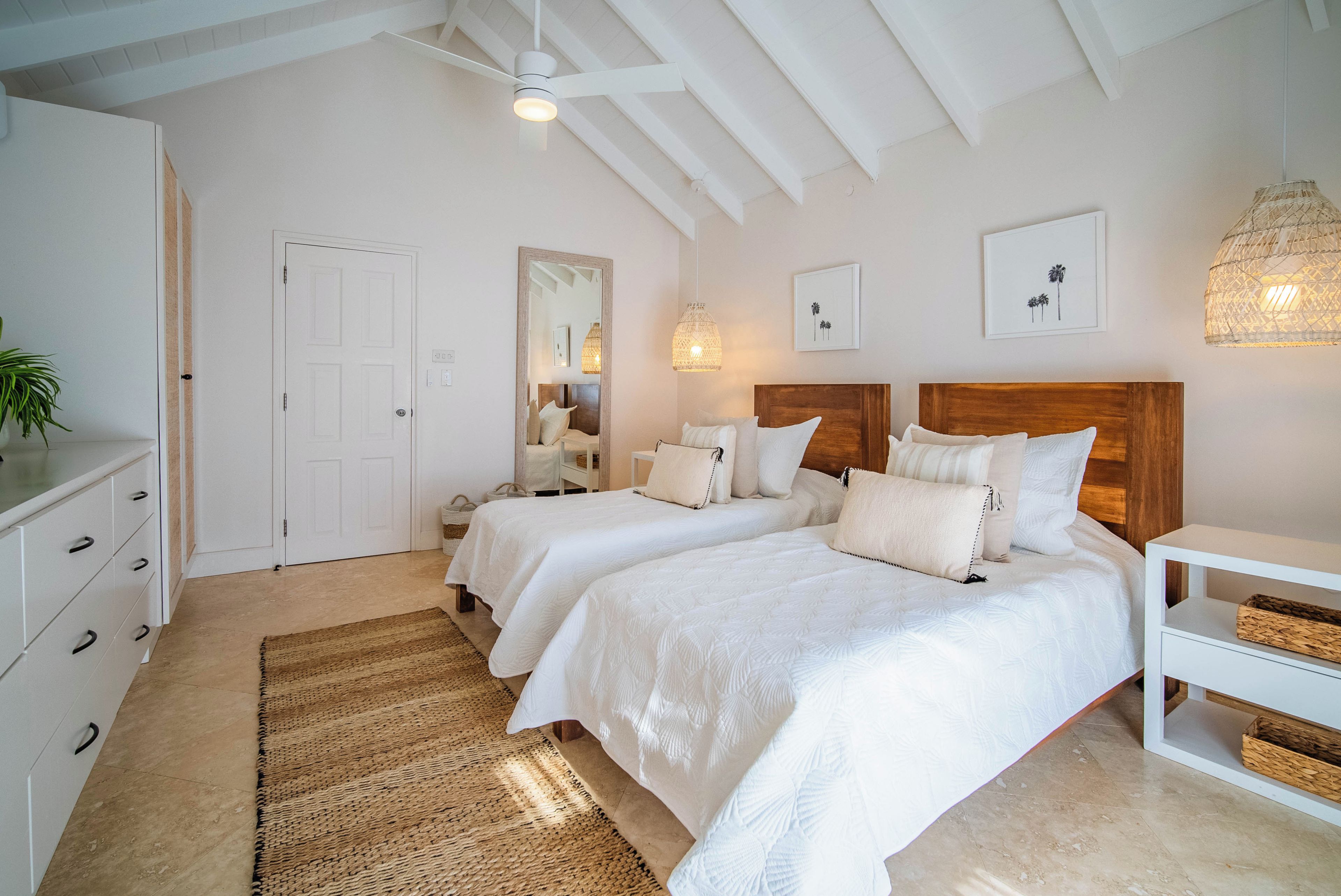 Ein Schlafzimmer mit zwei Betten, einem Holzkopfstück, einer weißen Kommode, einem gewebten Teppich und einem Deckenventilator.