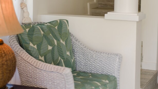 A living room with a wicker sofa, a green leaf-patterned pillow, a white curtain, and a staircase leading to an upper floor. - Little Good Harbour (Speightstown)