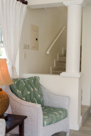 A living room with a wicker sofa, a green leaf-patterned pillow, a white curtain, and a staircase leading to an upper floor. - Little Good Harbour (Speightstown)