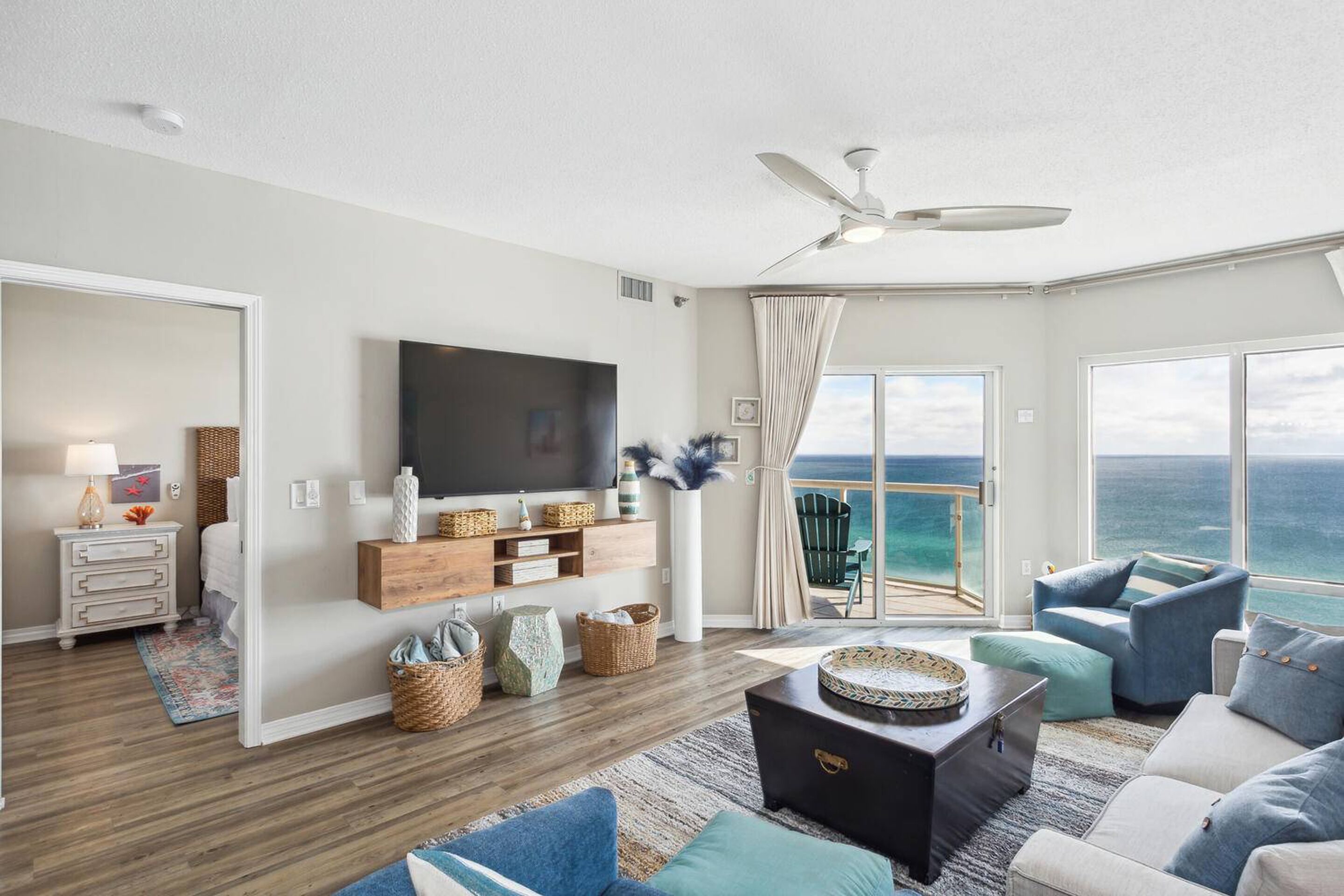 A spacious living room with a large TV, comfortable seating, and a balcony with a view of the ocean.