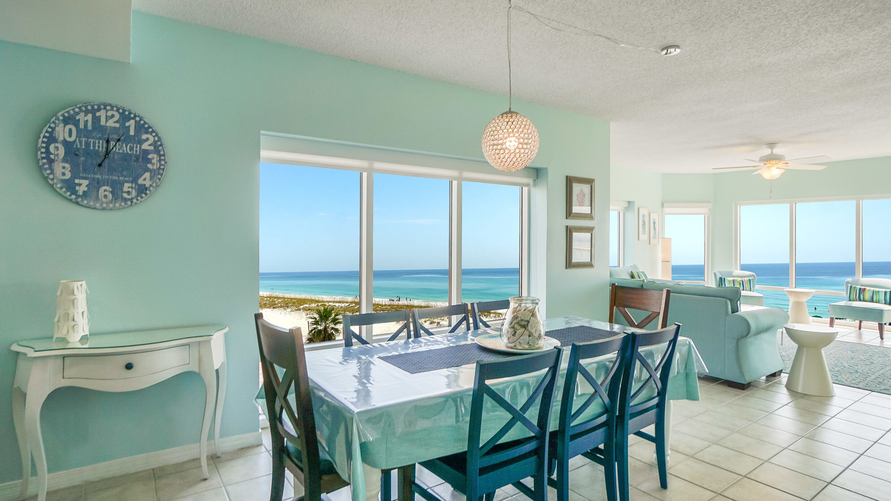 A dining area with a view of the beach, a clock on the wall, and a ceiling fan.
