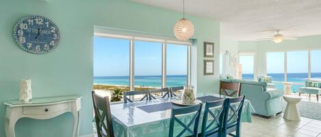 A dining area with a view of the beach, a clock on the wall, and a ceiling fan.