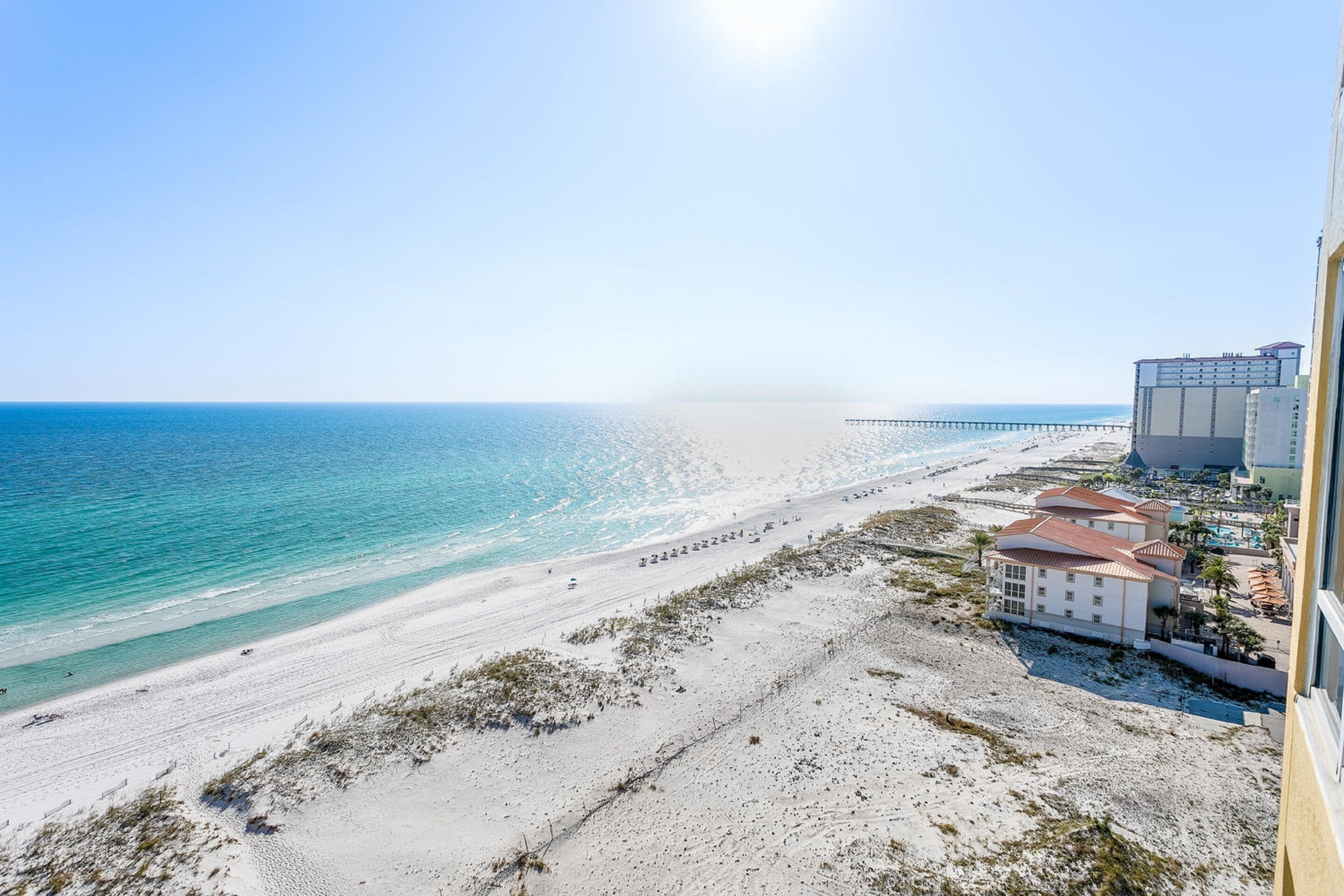 A beach view with a long pier extending into the ocean, a white building with a red roof, and a clear sky.