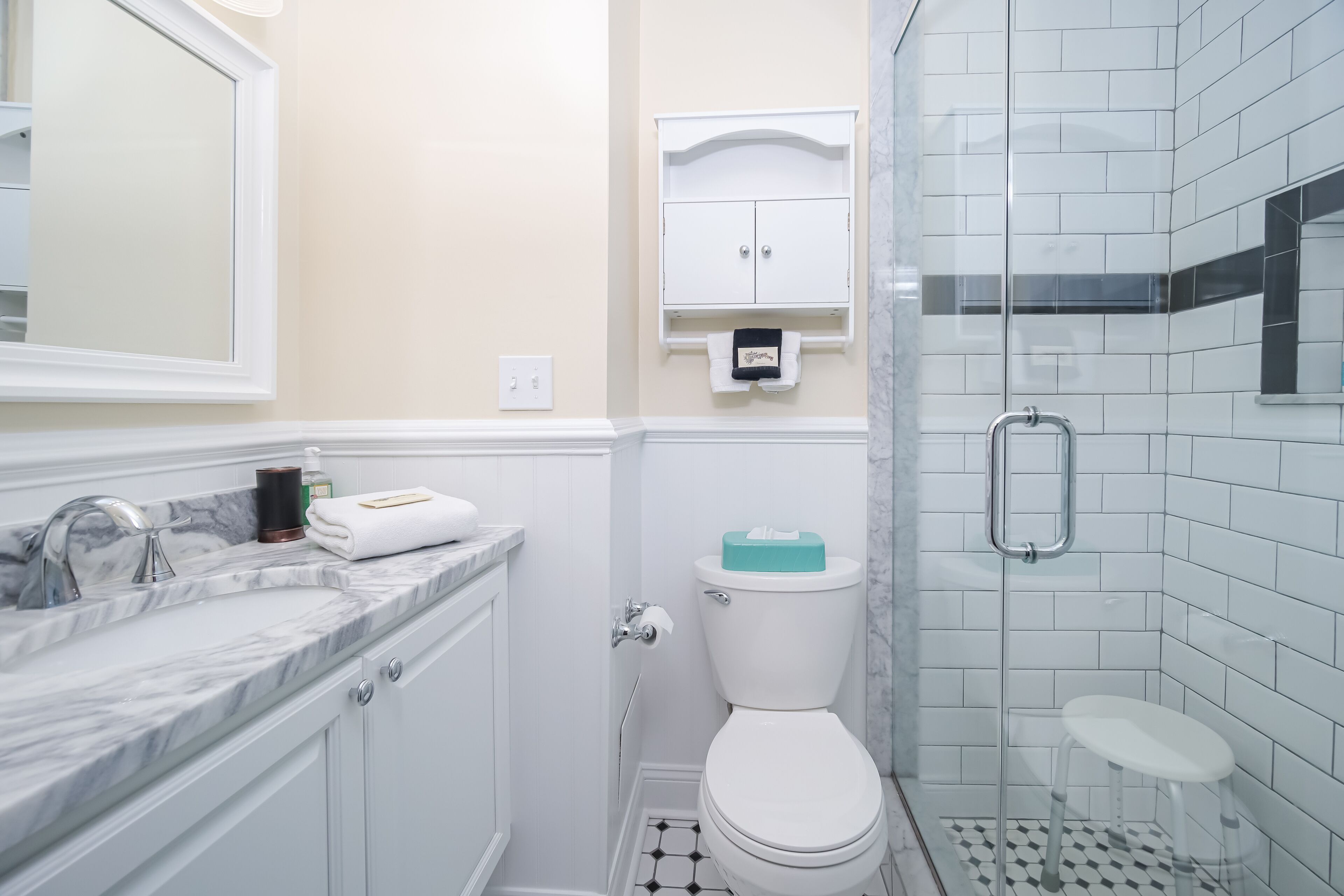 A bathroom with a white toilet, a marble sink, a glass shower enclosure, and a white cabinet with a towel dispenser.