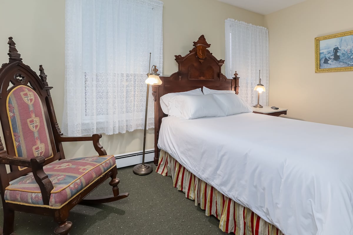 A bedroom with a wooden bed, a chair with a patterned cushion, a floor lamp, and a painting on the wall.