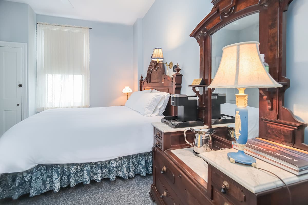 A neatly made bed with a white comforter and floral skirt, a wooden dresser with a mirror, a lamp, and a book titled "Coastal Journey" on top.