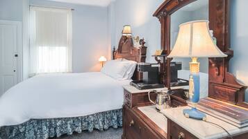 A neatly made bed with a white comforter and floral skirt, a wooden dresser with a mirror, a lamp, and a book titled "Coastal Journey" on top.