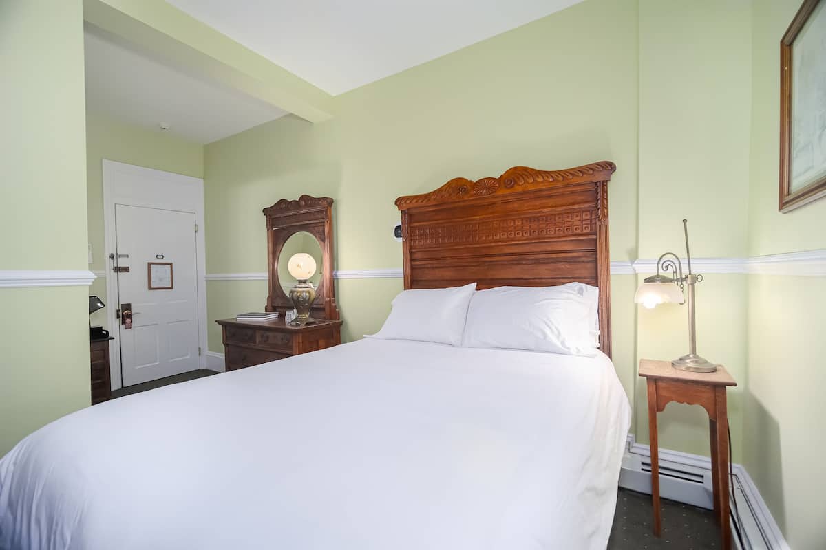 A neatly made bed in a room with a wooden headboard, a wooden nightstand with a lamp, a mirror, and a door.