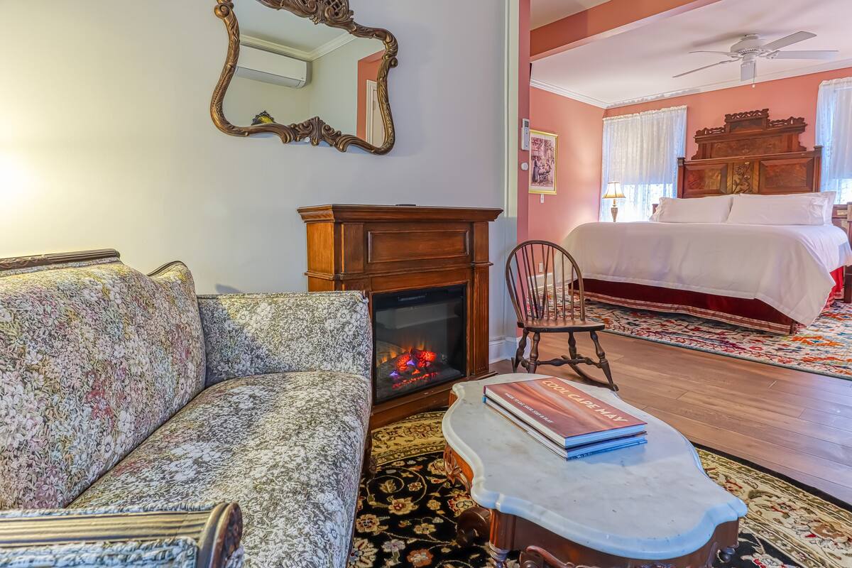 A cozy living room with a fireplace, a sofa, a coffee table with books, and a bed in the background.
