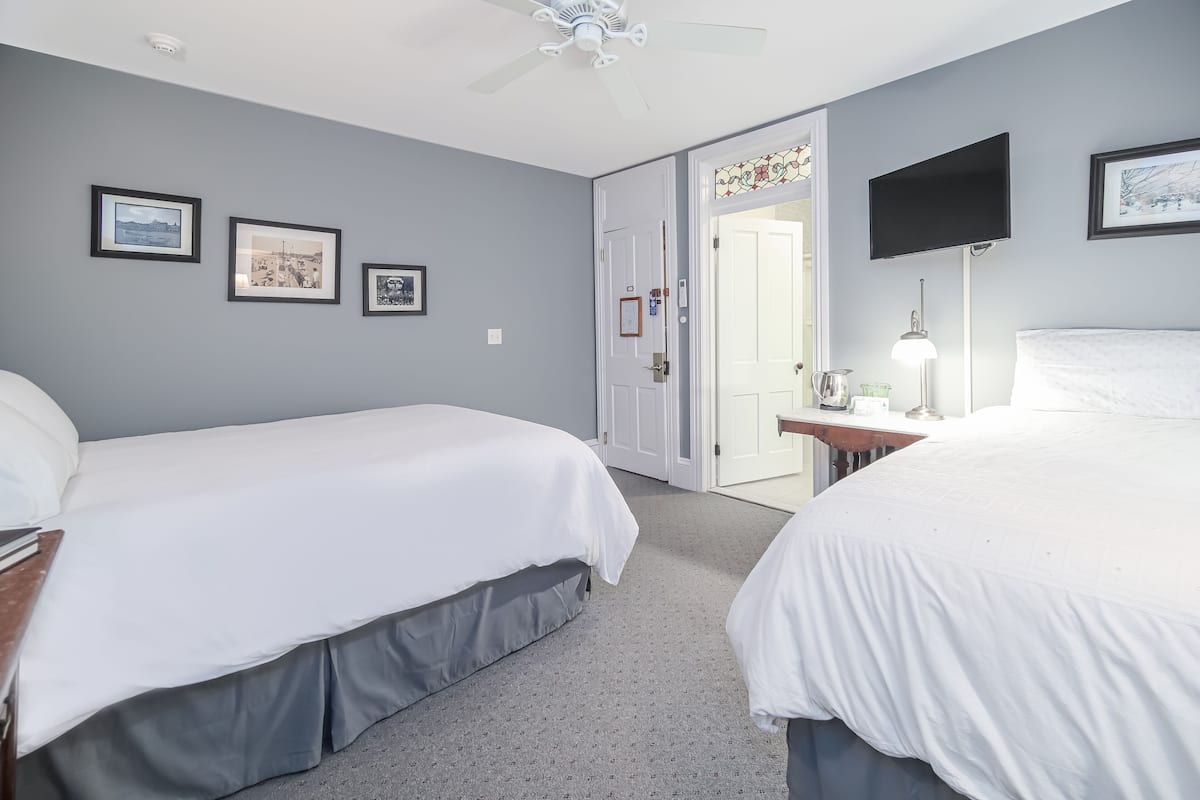 A hotel room with two beds, a ceiling fan, a TV, and framed pictures on the wall.
