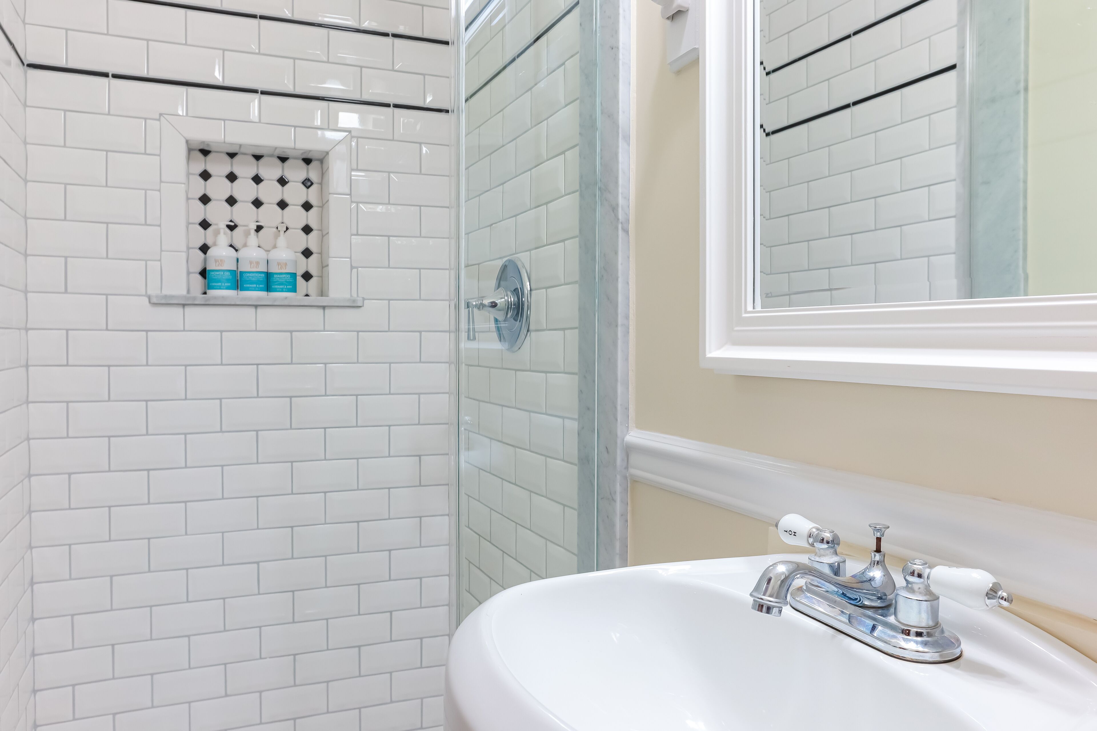 A bathroom with white tiled walls, a glass shower enclosure, and a white sink with chrome fixtures.