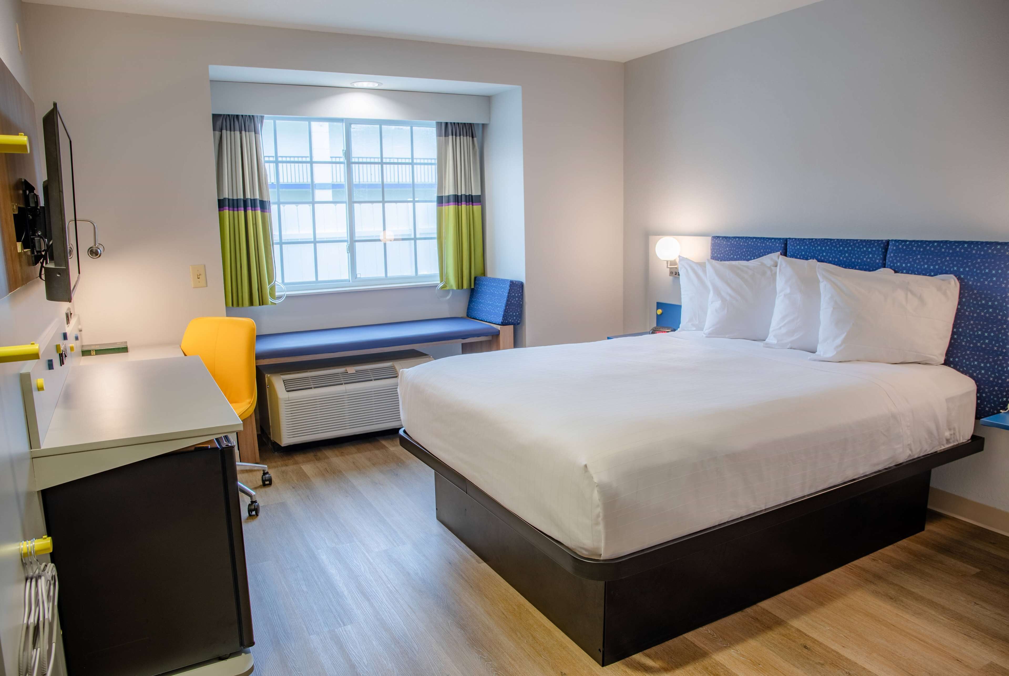 A hotel room with a bed, a desk, a chair, a window with curtains, and a heater.