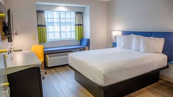 A hotel room with a bed, a desk, a chair, a window with curtains, and a heater.