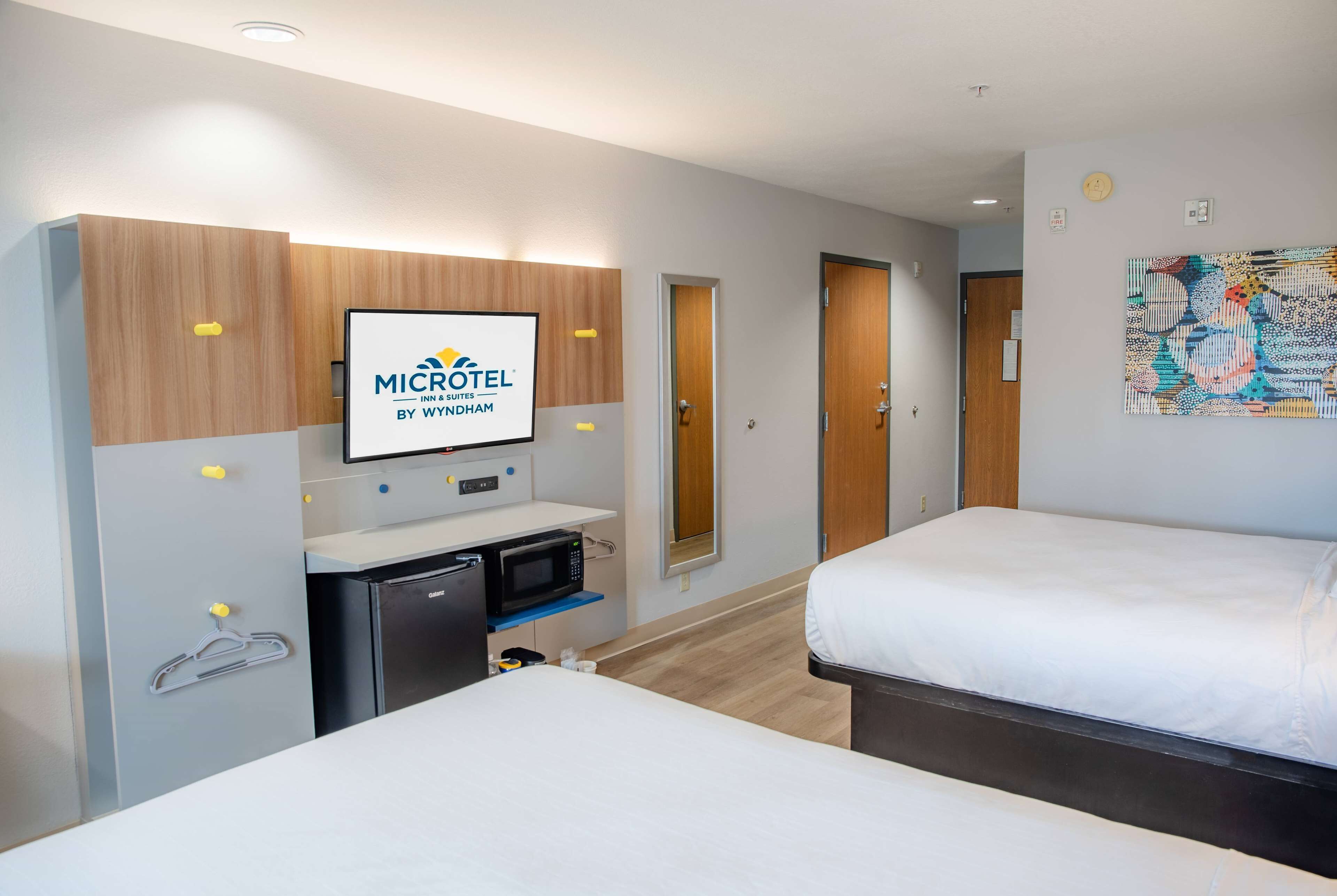 A hotel room with a bed, a desk with a microwave, a small fridge, and a TV displaying the hotel's logo.