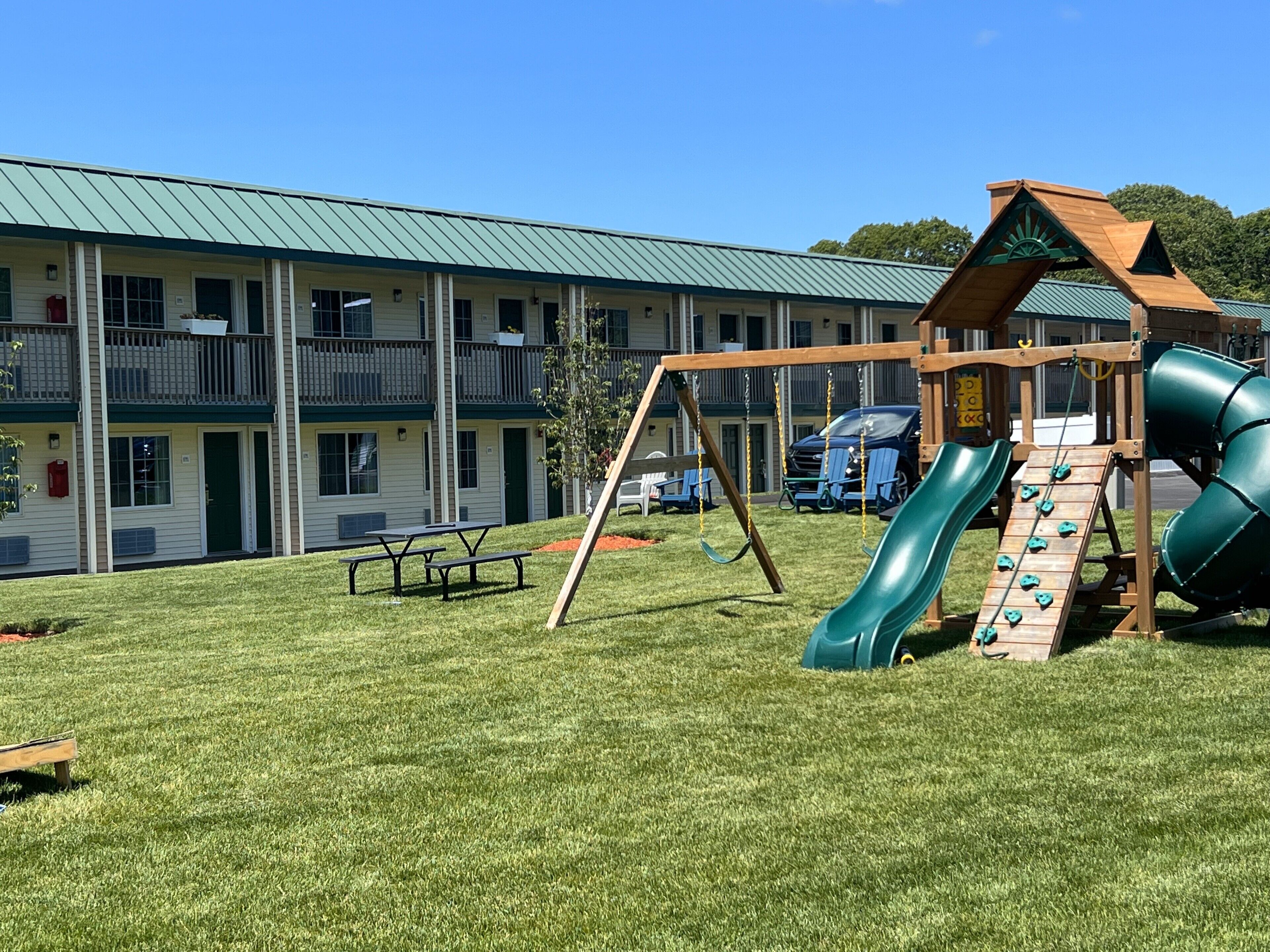 A playground with a slide and swings, a picnic table, and a building with a green roof and balconies in the background.