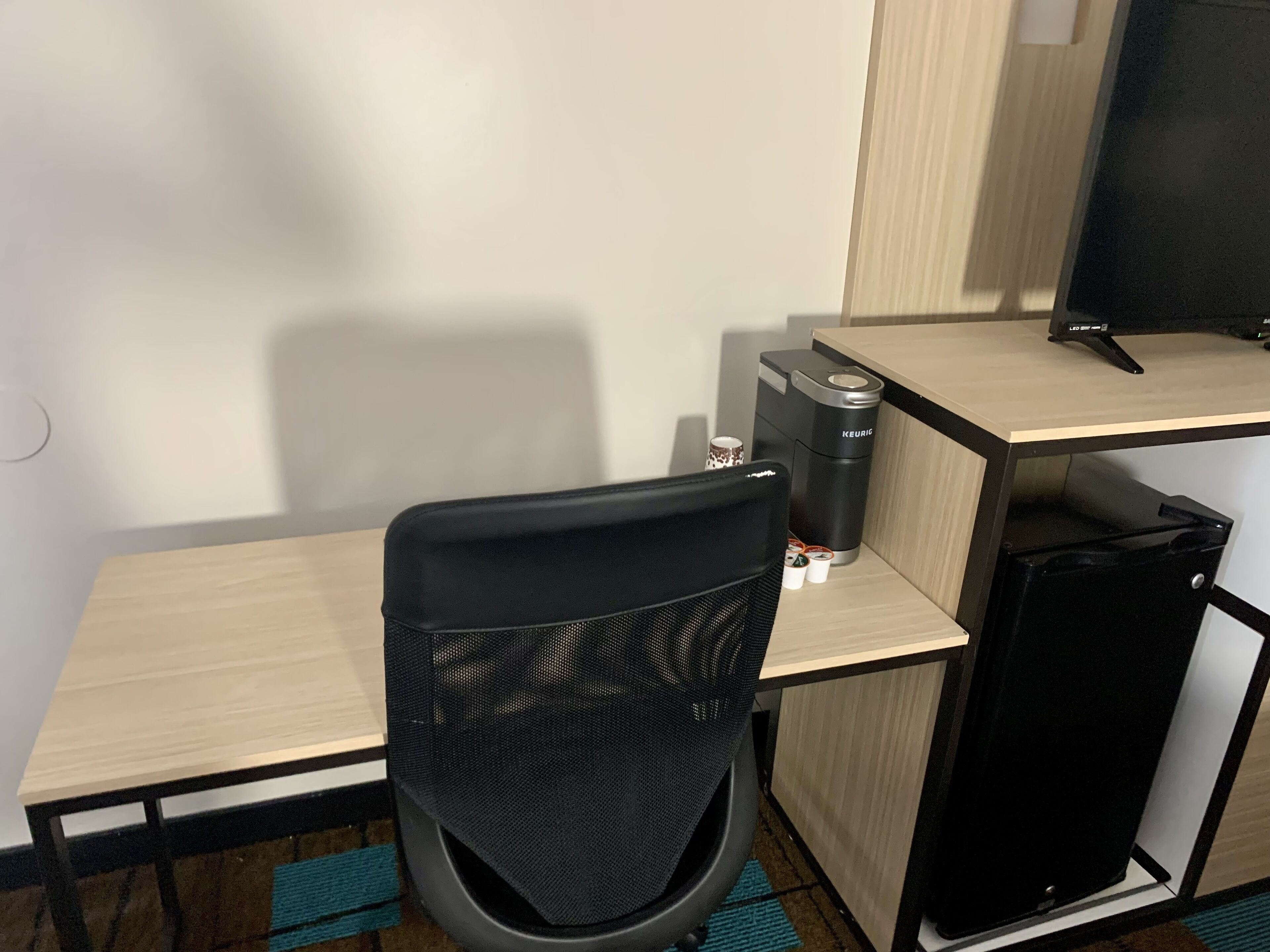 Een compacte hotelkamer met een enkel bureau, een zwarte bureaustoel, een koffiezetapparaat en een kleine koelkast.