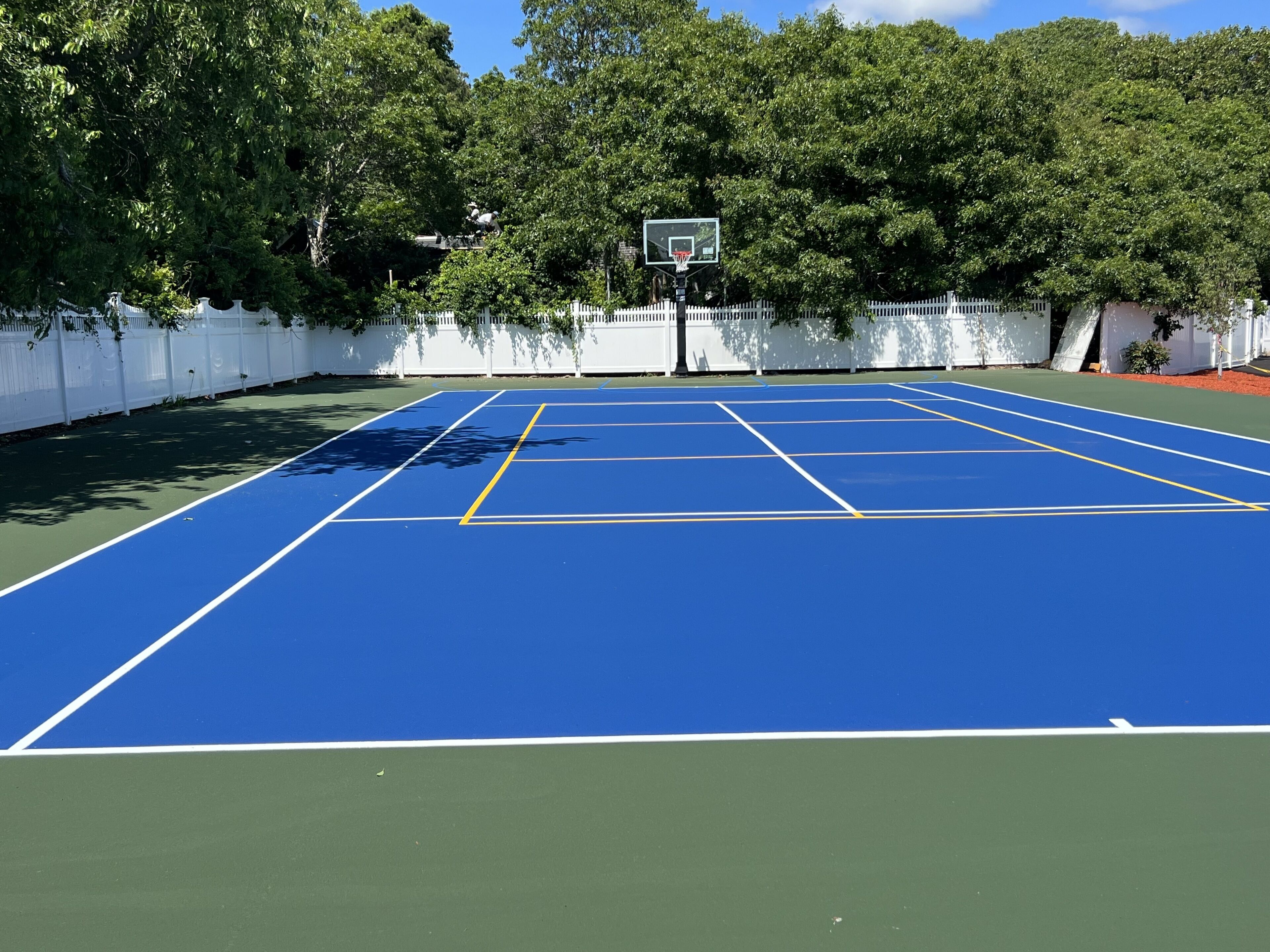 A blue tennis court with a basketball hoop, surrounded by a white fence and trees.