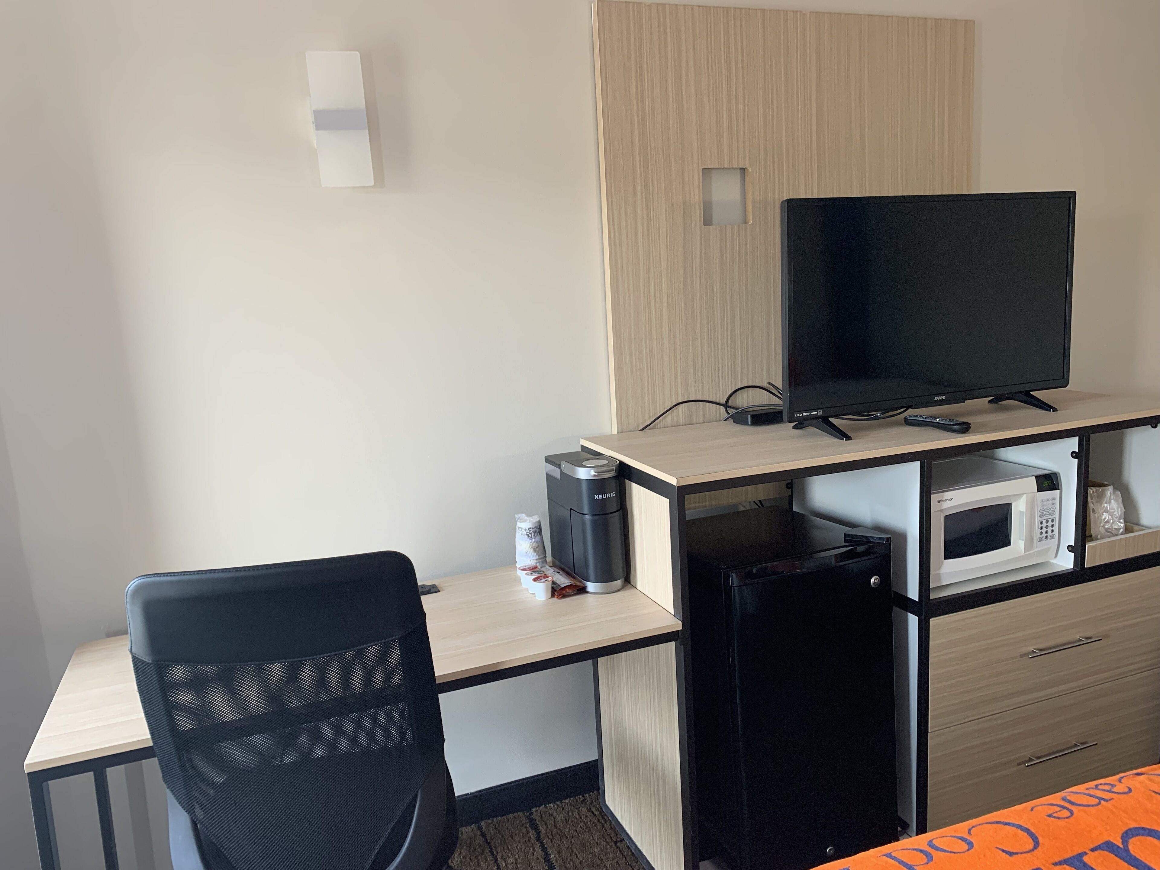 Quarto de hotel compacto com escrivaninha, cadeira de escritório preta, televisão, micro-ondas e cafeteira Keurig.