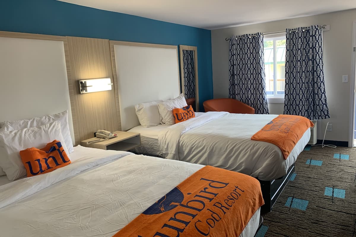 Une chambre d’hôtel avec deux lits, une table de chevet avec un téléphone, une chaise et une fenêtre avec des rideaux à motifs.