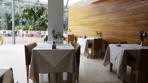An indoor dining area with tables set for two, wooden panel walls, and a view of greenery outside.