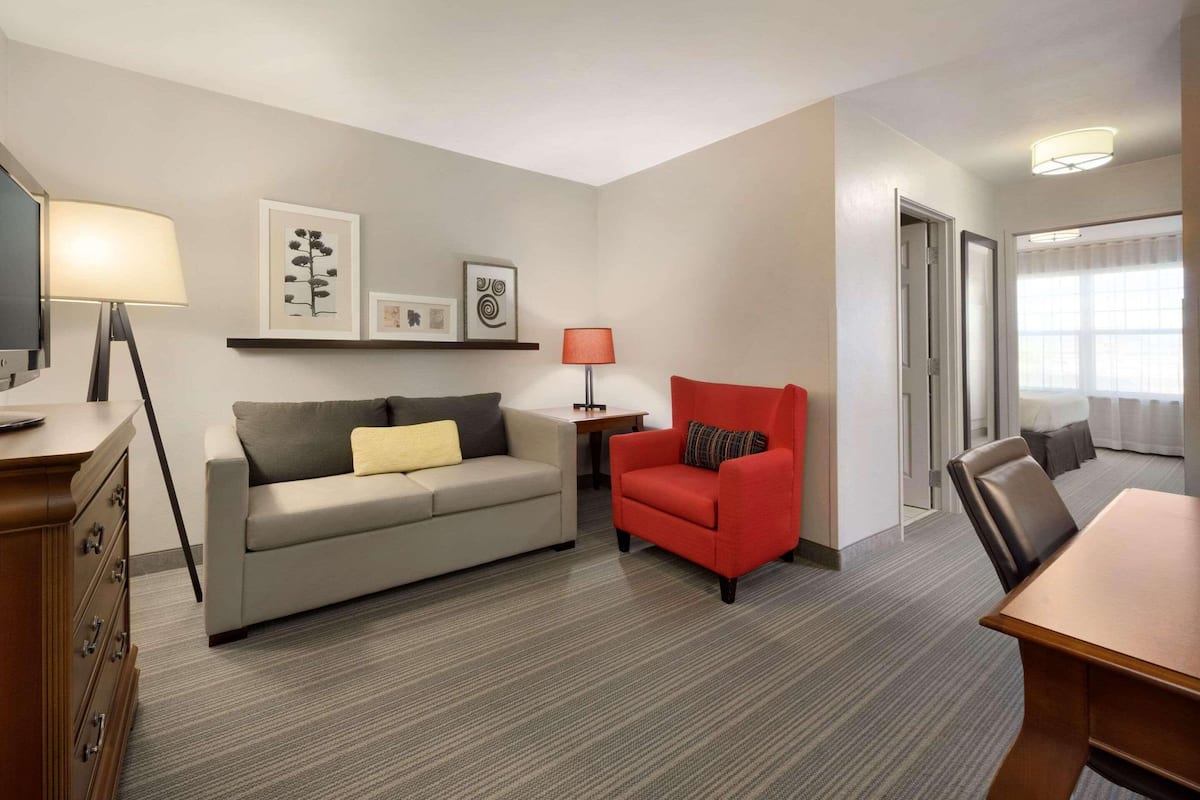 A hotel room with a sofa, a red armchair, a dining table, a lamp, and framed artwork on the wall.