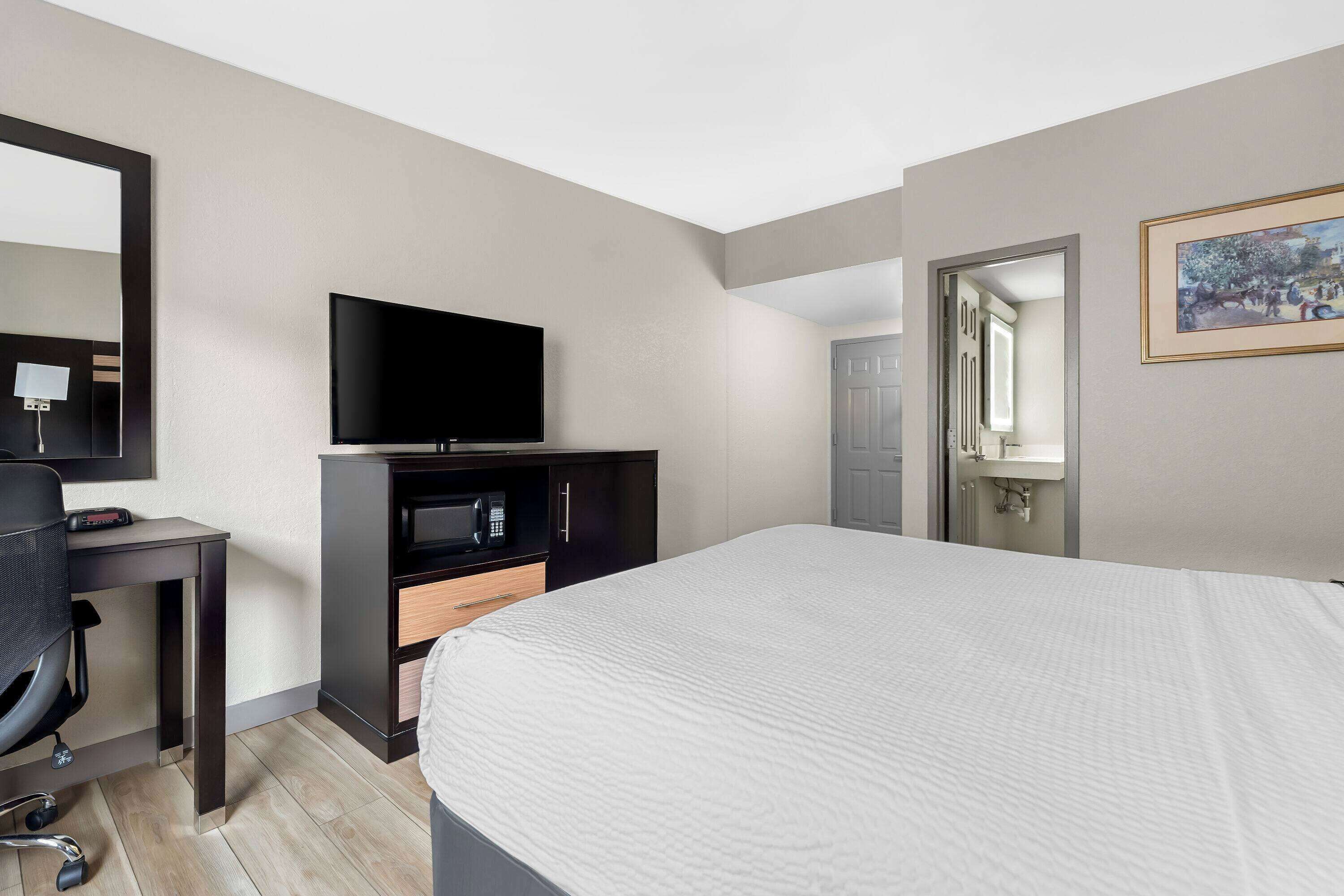 A modern hotel room with a large bed, a flat-screen TV, a desk with a chair, a microwave, and a door leading to a bathroom.