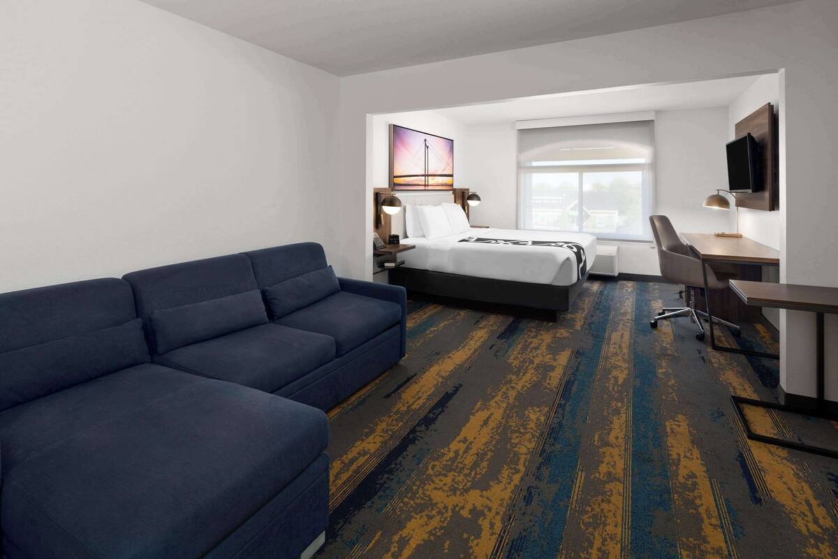 A hotel room with a large blue sofa, a bed with a white comforter, a desk with a chair, and a television mounted on the wall.