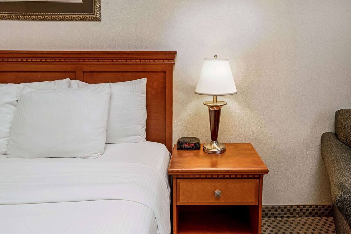 A hotel room with a wooden headboard, a bedside table with a lamp, a clock, and a chair.