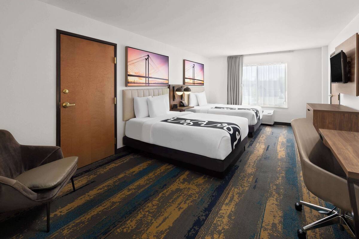 A hotel room with two beds, a desk, a chair, a TV, and artwork on the walls.